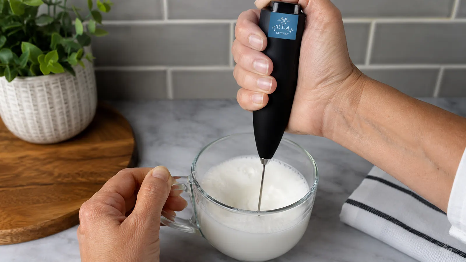 Hand holding a black handheld milk frother wand submerged in a glass mug, frothing milk