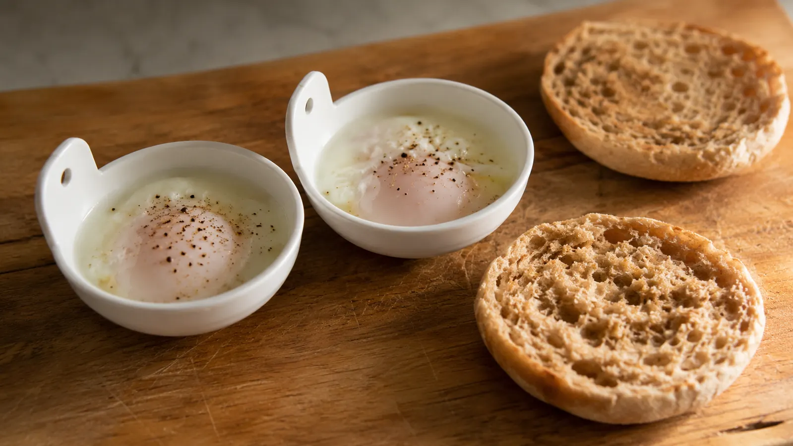 Freshly poached eggs in the small Dash poaching cups resting on the counter next to a whole-grain English muffin