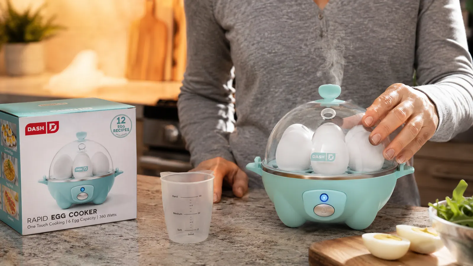 Close-up of the Dash egg cooker with steam rising and its measuring cup beside it on the counter