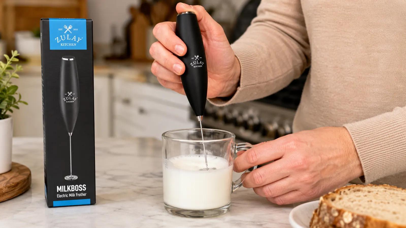 Hand holding the Zulay Kitchen milk frother wand above a glass mug, frothing steamed milk into a thick foam