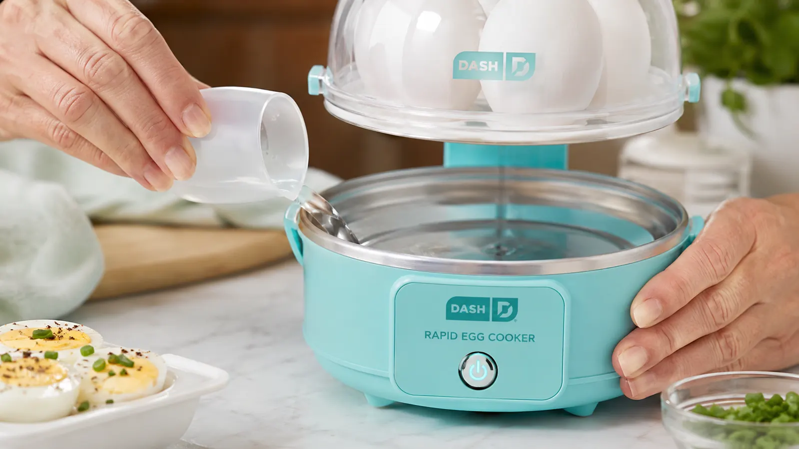 Hand using the included measuring cup to pour water into the Dash Rapid Egg Cooker base, eggs already loaded in the tray